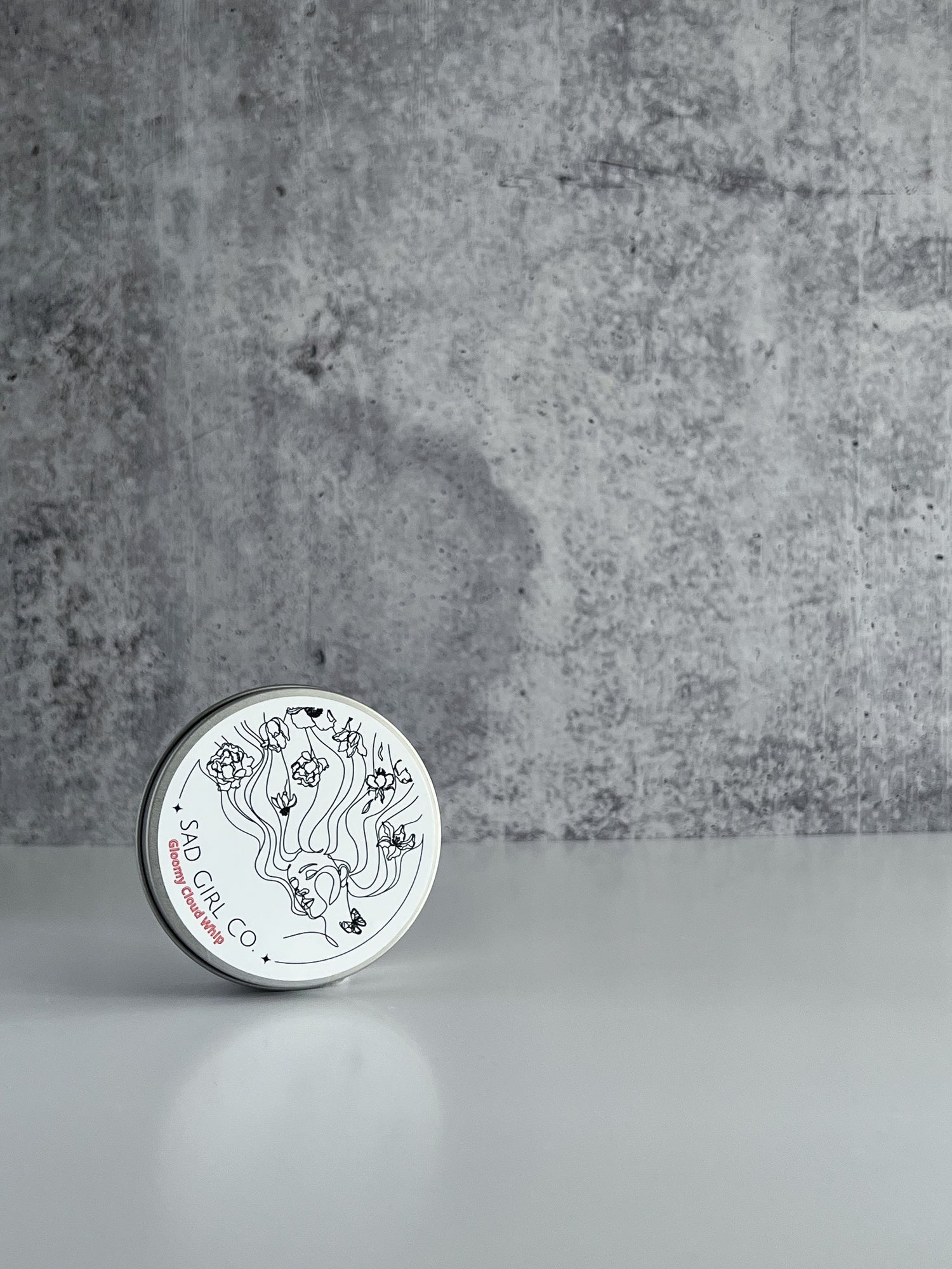 Silver tin sitting upright on a white surface with a cement background. Tin is labeled with Sad Girl Co. branding.